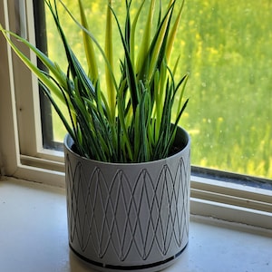 May include: A grey planter with a geometric design holds a green and yellow artificial plant. The planter is sitting on a windowsill.