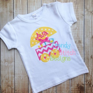 Puede incluir: Camiseta blanca con una sombrilla amarilla y blanca con lunares, un carrito de helados de chevron rosa y blanco, y el nombre "Ava" bordado en la sombrilla. El texto "Trendy Stitch Designs" también está bordado en la camiseta.