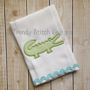 May include: White burp cloth with a green alligator applique and a blue scalloped edge. The alligator has a striped pattern and a blue eye. The text "Trendy Stitch Designs" is embroidered on the cloth.