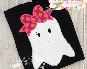 Puede incluir: Camiseta negra con un aplique de fantasma blanco adornado con un lazo rosa de lunares. El fantasma tiene una cara sonriente y pestañas negras. El diseño es de Trendy Stitch Designs.