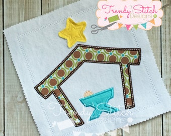 May include: An embroidered nativity scene on a white square. The scene includes a teal manger, a yellow star, and a brown and green patterned stable. The design is stitched on a white fabric with a decorative edge.