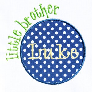 May include: A blue and white polka dot circle with a yellow embroidered name "Luke" inside. The circle is surrounded by a green embroidered phrase "little brother".