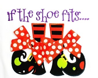 May include: White background with a Halloween-themed design. The image features a pair of black witch shoes with green accents, orange and white polka dot bows, and orange and black striped legs. The text "if the shoe fits..." is embroidered in purple.