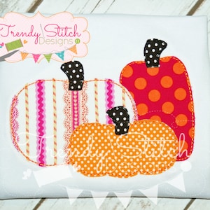 May include: White t-shirt with three appliqued pumpkins. The pumpkins feature various patterns, including stripes, polka dots, and lace trim. The pumpkins have black polka dot stems. The text "Trendy Stitch Designs" is visible.