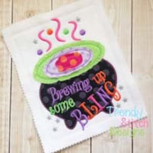 May include: A white fabric square featuring an embroidered black cauldron with a green rim and purple interior. The text reads Brewing up some BLING in purple, green, and orange. Pink swirls, colourful dots, and silver stars surround the cauldron.