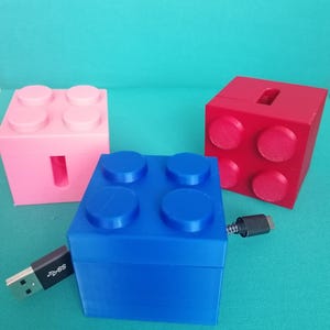 Könnte beinhalten: Drei bunte, blockförmige USB-Ladegeräte in Rosa, Blau und Rot. Jedes Ladegerät hat ein anderes Design, wobei das blaue mit einem USB-Kabel verbunden ist. Die Ladegeräte befinden sich auf einer türkisfarbenen Oberfläche.