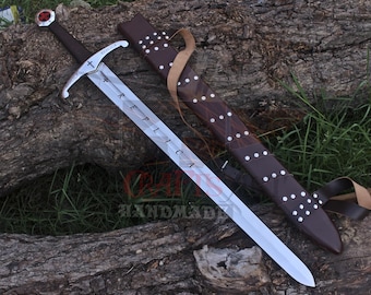 May include: A silver sword with a brown handle and a red gem at the pommel, resting on a log. The blade has the word "REPLICA" etched on it. A brown leather sheath with white dot accents is next to the sword.