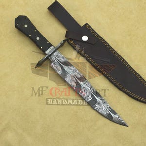 Può includere: Un pugnale fatto a mano con un manico in legno nero e una lama con un disegno a piume. Il pugnale è accompagnato da un fodero in pelle marrone. Il testo "MF Craft Handmade" è visibile.