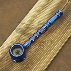 Puede incluir: Una pipa de metal azul con un patrón de panal en el tallo y una rejilla en el cuenco. La pipa está sobre una superficie de madera. La palabra "HANDMADE" es visible en la madera.