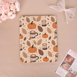 Pode incluir: Uma capa para iPad com um padrão de outono. O design inclui abóboras laranja, folhas castanhas e canecas de café num fundo bege. A capa tem formato retangular com uma capa dobrável.