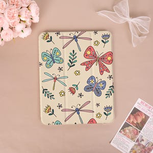 May include: A beige tablet case featuring a colorful pattern of butterflies, dragonflies, flowers, and leaves. The design includes blue, pink, yellow, and green elements. The case is rectangular and has a smooth surface.