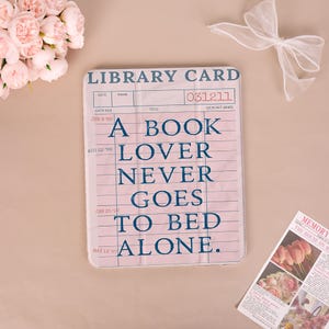 May include: A pink library card-themed case with the text "A BOOK LOVER NEVER GOES TO BED ALONE" in navy blue. The case has a vintage design with lines and dates, resembling a classic library card. A bow and flowers are in the background.