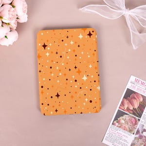 May include: An orange tablet case with a repeating pattern of white, brown, and orange stars and dots. The case is rectangular with rounded corners. The background is a light pink surface with flowers and a newspaper.