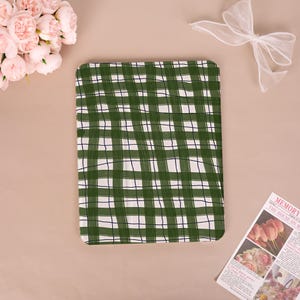 May include: A green and white checkered pattern fabric, possibly a tablecloth or blanket, dominates the image. The pattern is a grid of dark green lines on a white background. A bouquet of pink roses and a sheer white bow are in the background.