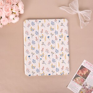 May include: White tablet case with a floral pattern featuring blue, yellow, and red sprigs. The case has a folding cover and a soft, matte finish. The design is a repeating pattern of stylized flowers and leaves.