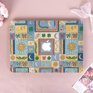 May include: A laptop skin featuring a repeating grid pattern of celestial and botanical motifs including yellow suns, crescent moons, green four-leaf clovers, red bows, and hands with star symbols in shades of blue, yellow, and red.