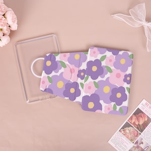 May include: A rectangular protective case for an electronic device featuring a floral pattern of purple and pink flowers with yellow centers and green leaves on a white background, accompanied by a separate clear plastic frame component.