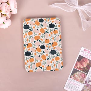 May include: A rectangular notebook with a white background and a repeating pattern of pumpkins in orange, black, and pink, with green leaves and orange accents. The notebook is on a pink surface.