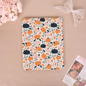 Peut inclure: Une housse de tablette blanche avec un motif répété de citrouilles orange, noires et roses, ainsi que des feuilles vertes et du feuillage orange. L'étui rectangulaire est conçu pour protéger une tablette.