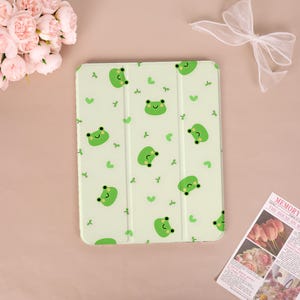 Op de afbeelding: Een lichtgroene tablethoes met een herhalend patroon van kleine, lachende cartoonkikkers en piepkleine groene blaadjes. De hoes heeft een drievoudig vouwontwerp met zichtbare verticale vouwen en een gladde, matte afwerking.