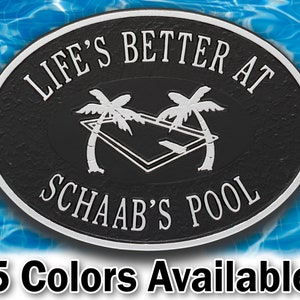 May include: Black oval-shaped metal sign with white text that reads "Life's Better at Schaab's Pool". The sign features two palm trees and a pool with a diving board.