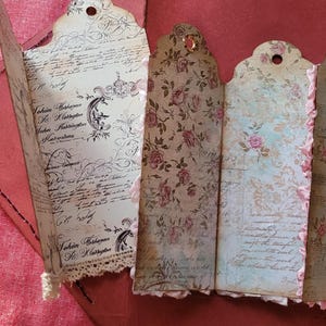 May include: A collection of vintage-style paper tags with decorative edges. The tags feature floral and script designs in shades of cream, pink, and brown. Some tags have lace trim and metal eyelets. The tags are arranged on a red surface.
