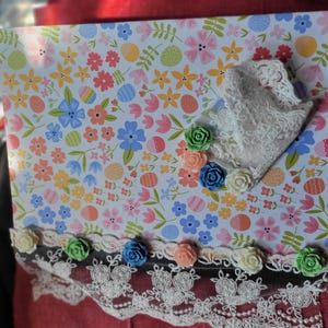 May include: A decorative floral-patterned notebook with a white lace trim and colorful rose embellishments. The notebook features a light blue background with various flowers and Easter egg designs. The trim is accented with small, colorful roses.