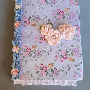 May include: A floral-patterned journal with a light blue background and pink roses. The journal is decorated with a blue ruffled ribbon and pink paper flowers. A white lace trim adorns the bottom edge.