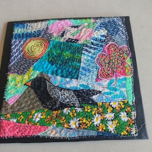 Könnte beinhalten: Ein quadratisches Textilkunstwerk mit einem schwarzen Vogel mit orangefarbenem Schnabel, vor dem Hintergrund bunter Stoffflicken. Das Kunstwerk umfasst eine gestickte Blume, eine Sonne und ein Gänseblümchenfeld. Das Werk ist mit einem schwarzen Rahmen versehen.