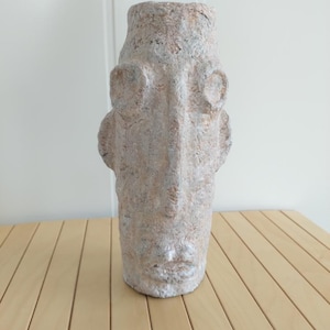 Op de afbeelding: Een hoge, getextureerde sculptuur van een menselijk gezicht. De sculptuur is voornamelijk beige met hints van grijs en roze. Het gezicht heeft prominente kenmerken, waaronder grote oogkassen en een duidelijke neus en mond. De sculptuur is waarschijnlijk een decoratief kunstwerk.