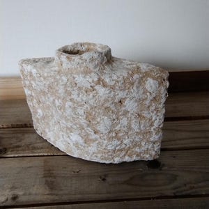 Op de afbeelding: Een ruwe, getextureerde, beige en witte keramische vaas met een brede, ronde opening. De vaas staat op een houten oppervlak.