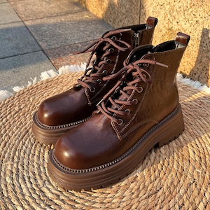 Puede incluir: Un par de botines de cuero marrón con suelas gruesas y gruesas. Las botas tienen un diseño con cordones con cordones marrones y una cremallera lateral. Las botas están colocadas sobre una estera redonda tejida.