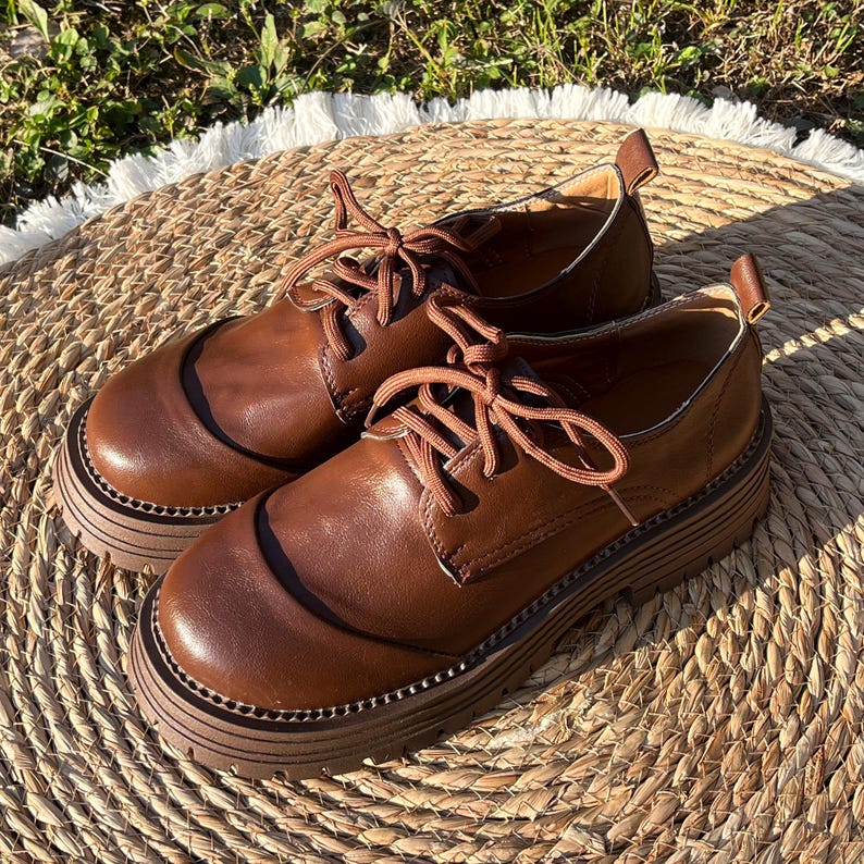 May include: A pair of brown leather oxford shoes with thick, ridged soles. The shoes have a classic lace-up design with brown laces and a rounded toe. The shoes are set on a woven, circular mat.