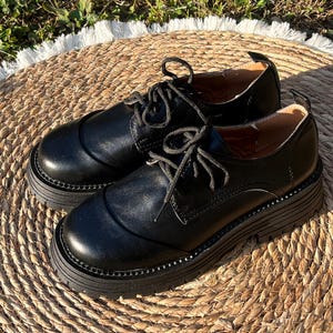 May include: A pair of black leather oxford shoes with thick, lug soles. The shoes have a classic lace-up design and a rounded toe. The shoes are sitting on a woven, circular mat.