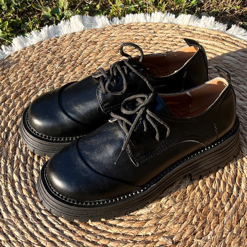 May include: A pair of black leather oxford shoes with thick, lug soles. The shoes have a classic lace-up design and a rounded toe. The shoes are sitting on a woven, circular mat.