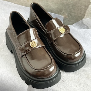 May include: A pair of brown leather loafers with a glossy finish. Each shoe features a gold-colored floral accent on the strap and a thick black sole. The shoes have a classic, preppy style.