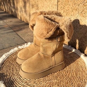 Puede incluir: Un par de botas de ante color canela con ribete de piel sintética. Las botas tienen un detalle de hebilla y una suela de plataforma gruesa. Las botas están sobre una estera tejida.