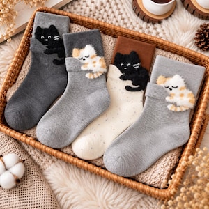 Könnte beinhalten: Vier Paar Socken mit flauschigen Katzenapplikationen. Die erste ist dunkelgrau mit einer schwarzen Katze, die zweite hellgrau mit einer weiß-orangen Katze, die dritte cremefarben mit einer schwarzen Katze und die vierte hellgrau mit einer weiß-orangen Katze.