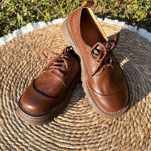 May include: A pair of brown leather oxford shoes with thick, ridged soles. The shoes have a classic lace-up design and a rounded toe. The leather has a polished finish, and the shoes are set on a woven mat.