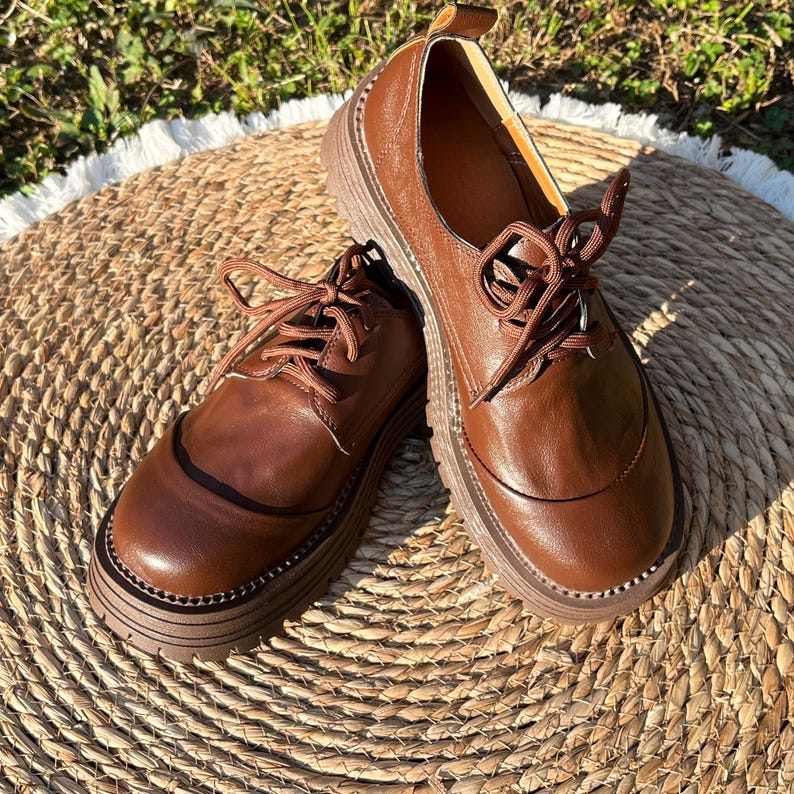 May include: A pair of brown leather oxford shoes with thick, ridged soles. The shoes have a classic lace-up design and a rounded toe. The leather has a polished finish, and the shoes are set on a woven mat.