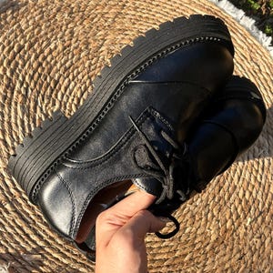 May include: Black leather oxford shoes with thick, ridged soles. The shoes have a classic lace-up design and a stitched welt. The image shows the shoes on a woven mat, highlighting their texture and style. A hand is holding the shoe.