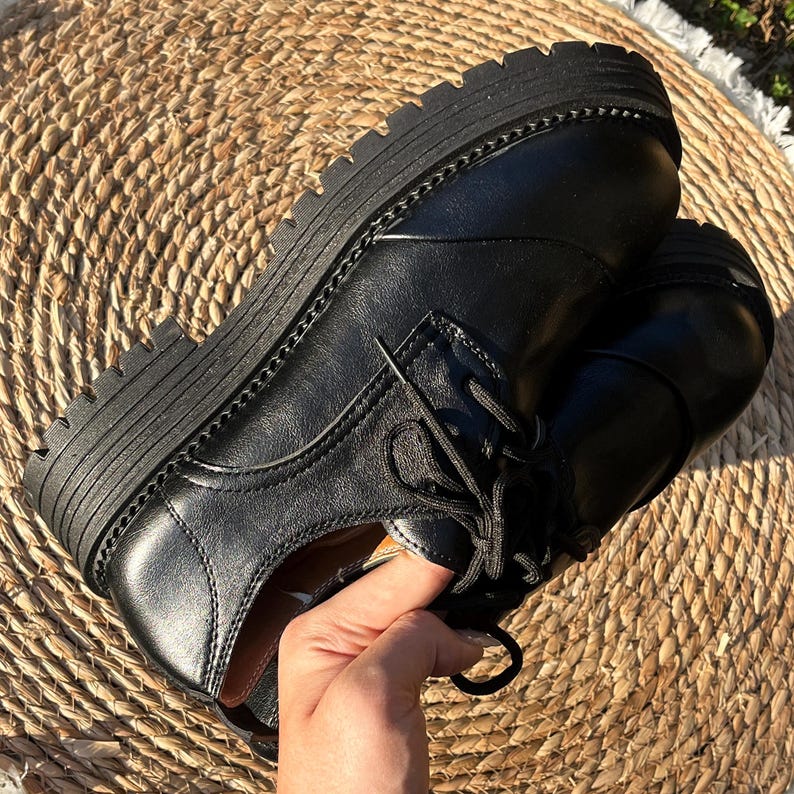 May include: Black leather oxford shoes with thick, ridged soles. The shoes have a classic lace-up design and a stitched welt. The image shows the shoes on a woven mat, highlighting their texture and style. A hand is holding the shoe.