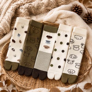 May include: Five pairs of split-toe socks featuring various designs including small dog faces, polka dots, and horizontal stripes in shades of cream, olive green, and dark brown. Each pair has a distinct toe separation and ribbed or smooth fabric.