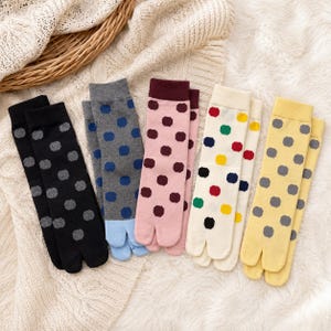May include: Five pairs of split-toe socks featuring polka dot patterns in black, gray with light blue toes, pink with maroon cuffs, cream with multicolored dots, and yellow. Each pair has a distinct color scheme and a separated big toe design.