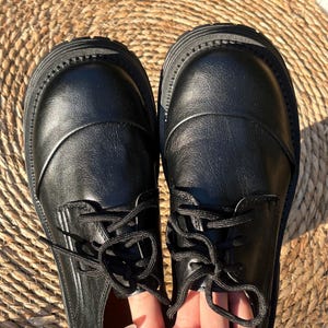 May include: A pair of black leather shoes with thick soles and black laces. The shoes have a rounded toe and a smooth, polished finish. The laces are tied in a neat bow.
