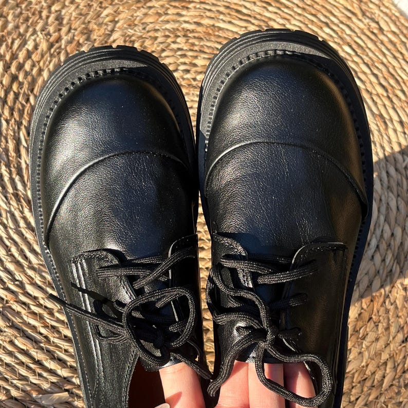 May include: A pair of black leather shoes with thick soles and black laces. The shoes have a rounded toe and a smooth, polished finish. The laces are tied in a neat bow.