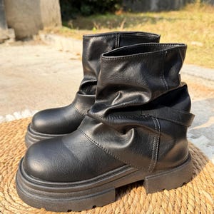 May include: A pair of black ankle boots with a chunky platform sole. The boots have a scrunched design at the top and a strap detail. The soles are approximately 2 inches high.
