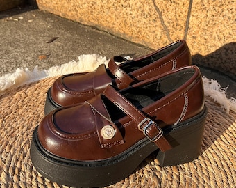 May include: A pair of brown leather Mary Jane shoes with a chunky black platform sole. The shoes have a strap with a buckle closure and a decorative button. The shoes are sitting on a woven mat.