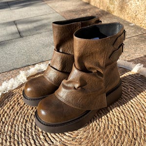 May include: A pair of brown leather ankle boots with a unique design. The boots feature a rounded toe, a thick sole, and decorative buckles. The leather has a textured appearance, and the boots are set on a woven mat.