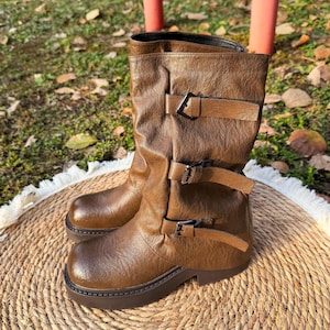 Puede incluir: Botas de cuero marrón con tres correas con hebilla. Las botas tienen una puntera redondeada y una suela gruesa. Las botas están sobre una estera tejida. Las botas son de color marrón medio.